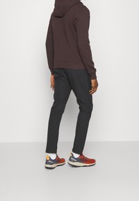 Felpa marrone scuro con tasca a marsupio, abbinata a pantaloni neri e sneaker rosse con accenti blu e gialli.