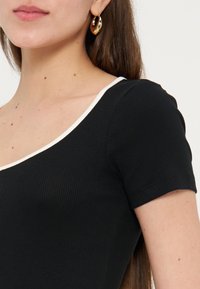 Γυναίκα που φοράει μαύρο κοντομάνικο ριμπ τοπ με λευκή επένδυση και χρυσό κρίκο σκουλαρίκι, εμφανίζοντας το λαιμό και την περιοχή των ώμων.