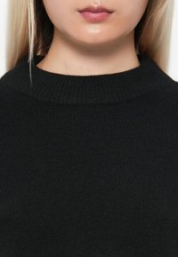 Fekete kötött pulóver magas, bordázott nyakkal. A mellkasi területről készült közeli felvételen látható a puha, texturált anyag.