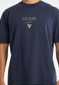 Man in een marineblauwe t-shirt met korte mouwen met de tekst "GUESS originals" en een klein driehoekig logo op de borst.