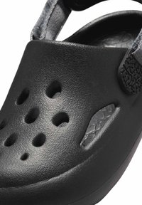 Svart gummiklump med ventilationshål, texturerad yta och tjock sula. Justerbar rem för en säker passform. Tjockt mönster för bra grepp.