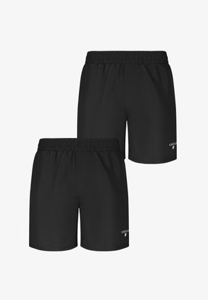 Homme musclé avec de nombreux tatouages, y compris un grand dessin de visage sur le torse, portant un short noir, debout pieds nus devant un fond gris uni.