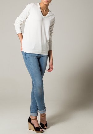 Witte V-hals trui met lange mouwen, gecombineerd met lichtblauwe, smal gesneden jeans. Model draagt zwarte wedge sandalen met enkelbandjes.