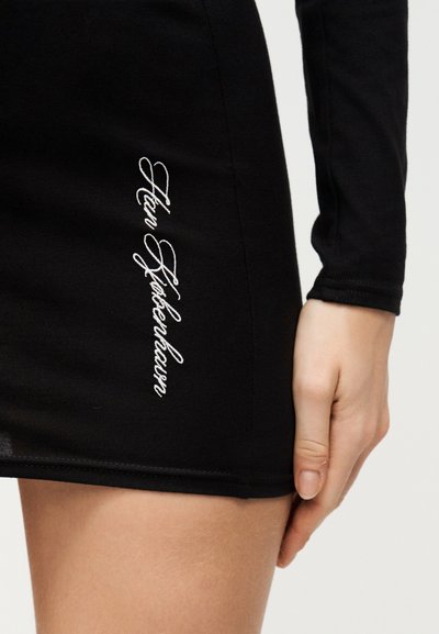 Κοντινό πλάνο ενός ατόμου που φοράει μαύρο ρούχο με λευκή καλλιγραφική επιγραφή «Tom Fröhkerbaum» κεντημένη κάθετα κοντά στον μηρό, χέρι που ξεκουράζεται δίπλα του.