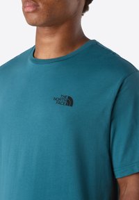 Hombre vistiendo una camiseta de cuello redondo de manga corta en color teal con el pequeño logo negro "The North Face" en el lado izquierdo del pecho.
