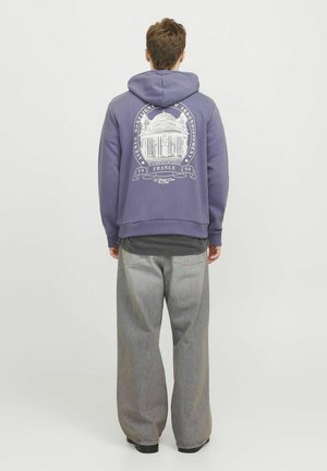 Fioletowy sweatshirt z kapturem z białym nadrukiem graficznym na plecach, przedstawiającym projekt architektoniczny i tekst. W połączeniu z szarymi spodniami typu wide-leg.