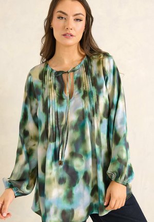 Kobieta ubrana w luźną bluzkę z długim rękawem, ozdobioną wzorem przypominającym akwarelę w odcieniach zieleni, niebieskiego i czarnego, z wiązaniem przy dekolcie.