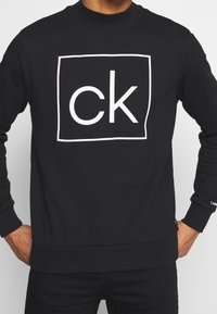 Hombre con sudadera negra con el logo blanco "ck" dentro de un cuadrado en el pecho, con las manos en las caderas, visible de cuello a caderas.
