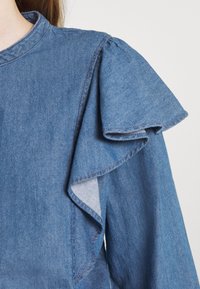 Denimbluse mit rundem Kragen, gepufften Ärmeln und geschichteten Rüschendetails an der Schulter, in einem hellblauen Farbton mit sichtbaren Nähten.