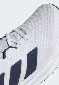 Gros plan d'une chaussure de sport blanche avec texture en maille, lacets blancs et trois bandes bleu foncé sur le côté.
