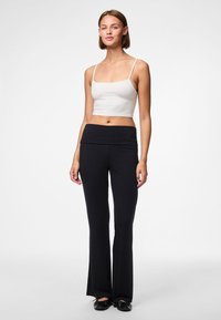 Vrouw staand in zwarte hoge taillebroek en wit kort tanktopje met dunne bandjes, tegen een effen witte achtergrond.