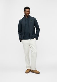 Νεαρός άνδρας στέκεται με τα χέρια στις τσέπες, φορώντας ναυτικό μπουφάν, σκούρο ριγέ πουκάμισο, ανοιχτόχρωμο παντελόνι και καφέ σουέντ παπούτσια σε λευκό φόντο.