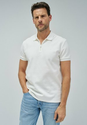 Camisa polo blanca con cuello con cremallera, de mangas cortas y tejido texturizado. Combinada con vaqueros de un azul claro, llevada de manera casual con las manos en los bolsillos.