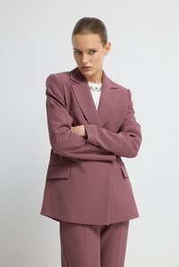 Blazer mauve clair cintré avec larges revers, deux poches avant, et une texture lisse, accompagné d'un haut blanc orné d'accents en cristal.
