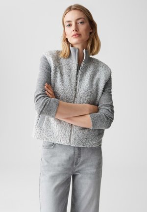 Donna con capelli biondi che indossa un maglione grigio con cerniera e trama e jeans grigio chiaro, in piedi con le braccia incrociate davanti a uno sfondo semplice.