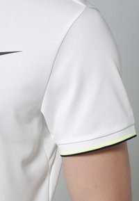 Biała koszulka sportowa z fakturowanej tkaniny, z czarnym logo Nike oraz kontrastującymi żółtymi i czarnymi wstawkami na rękawach.