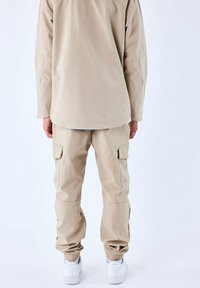 Beige waterdichte jas met geribbelde nek en manchester, gecombineerd met bijpassende cargobroek met zakken en elastische manchester, gedragen met witte sneakers.