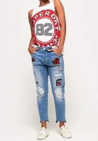 Vit ärmlös linne med röd cirkulär logotyp och svart text, tillsammans med ljusblå slitna jeans med färgglada lappar och vita sneakers.