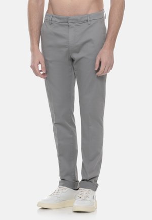 Uomo che indossa pantaloni grigi aderenti risvoltati alle caviglie e sneakers bianche, in piedi davanti a uno sfondo neutro.