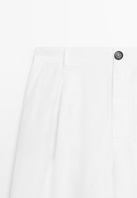 Bílé lněné kalhoty s plisovaným předním dílem, bočními kapsami a černým knoflíkem na pásu. Hladká textura a přiléhavý střih.