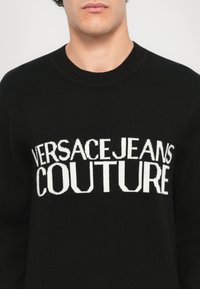 Črn pleten pulover z izpostavljenim belim logotipom "VERSACE JEANS COUTURE" v izrazitem črkem in klasičnim krojem izreza.