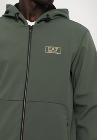 Giacca softshell verde con chiusura a zip, cappuccio regolabile e dettagli neri. Presenta un logo EA7 dorato sul petto. Texture liscia.