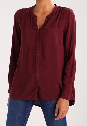 Vrouw in een bordeauxrode blouse met lange mouwen en knopen, voorzien van twee borstzakken, en blauwe jeans, poseert tegen een effen achtergrond.