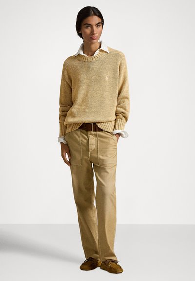 Bež pulover iz pletenine z rebrastimi manšetami, nošen preko bele srajce z ovratnikom. Kombiniran s svetlo rjavimi hlačami in rjavimi loaferji.