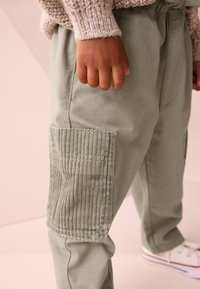 Pantaloni cargo verde chiaro con dettagli in tessuto a coste sulle tasche. Il materiale è liscio, con vita elastica e dettagli di cuciture.