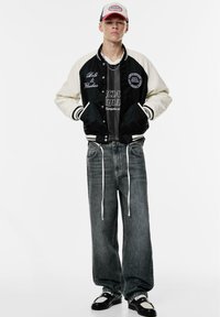Veste varsity noire et crème avec des patches brodés, t-shirt graphique gris, jean gris ample, et chaussures noires avec accents blancs.
