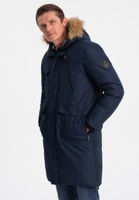 Námořnický modrý parka kabát s kožichem na kapuci, kapsami na zip a knoflíky, hladkou texturou a logem na rukávu.