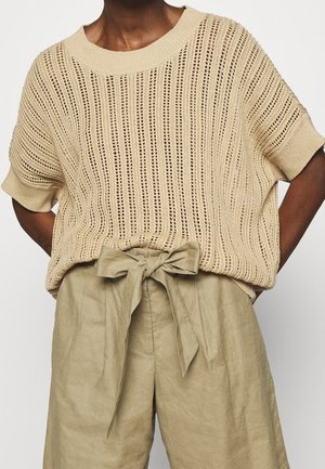 Beige knitted short-sleeve sweater with a loose fit, featuring an open weave pattern. Paired with light beige trousers tied at the waist.