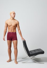 Bóxer burdeos con cintura elástica, acompañados de una silla de descanso negra minimalista con un marco de cromo y asiento acolchado.