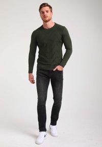 Maglione lavorato a maglia verde scuro con motivi geometrici testurizzati, abbinato a jeans grigi scuri e sneakers bianche. Il modello si trova su uno sfondo neutro.