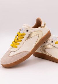Sneakers crema e beige con dettagli dorati, tomaia testurizzata, lacci gialli e suola in gomma. Presentano un design curvo e un'etichetta con logo.