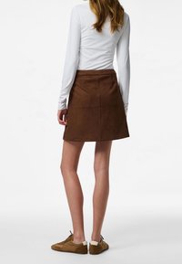 Femme portant un haut blanc à manches longues, une jupe marron jusqu'aux genoux et des baskets marron, se tenant de dos face à l'observateur sur un fond blanc.