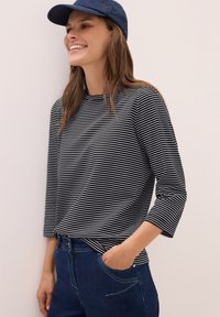 Leende kvinna iförd en marineblå keps, svartvit randig långärmad skjorta och mörkblå jeans med en hand i fickan, lutande mot en ljus vägg.