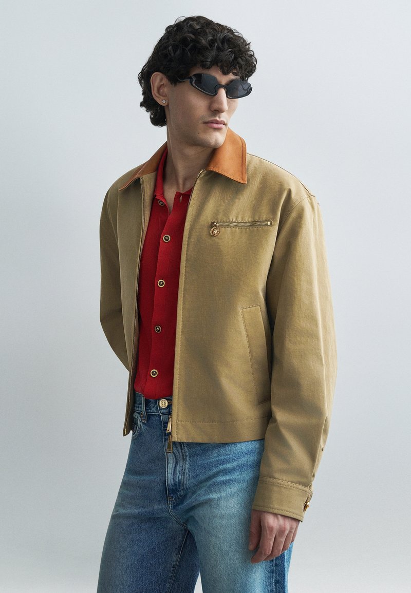 Vyras su garbanotais plaukais, dėvi juodus siaurus akinius nuo saulės, rudos spalvos striukę su odiniu apykakle, raudoną marškinių tipo palaidinę ir mėlynas džinsines kelnes, žvelgia į šoną.