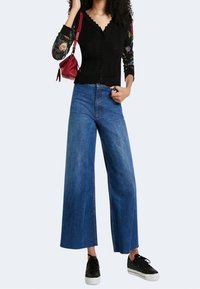 Musta ribbineulosneuletakki, jossa on reunuksellinen v-pääntie, yhdistetty leveälahkeisiin sinisiin farkkuihin ja mustiin lenkkareihin. Malli pitää punaisen käsilaukun.