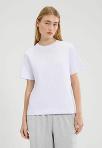 Jonge vrouw met lang blond haar draagt een eenvoudige witte t-shirt met korte mouwen en lichtgrijze broek, staand tegen een effen achtergrond.
