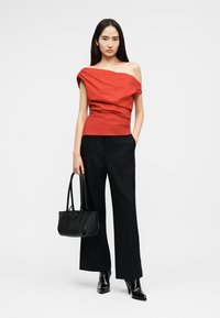 Γυναίκα που φοράει κόκκινο ασύμμετρο τοπ με έναν ώμο, μαύρο παντελόνι φαρδύ, μαύρες μπότες με τακούνι, κρατώντας μια μαύρη τσάντα.
