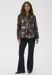 Donna in piedi che indossa una camicia a maniche lunghe con modello camouflage bottonata, jeans scuri a zampa d'elefante e scarpe nere a punta su uno sfondo bianco.