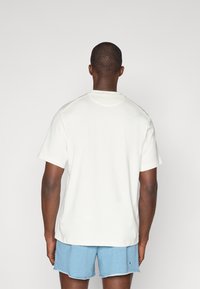 Camiseta blanca de manga corta con escote redondeado, textura suave y diseño minimalista, combinada con pantalones cortos azules.