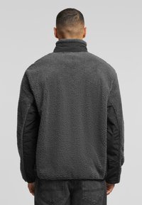 Gray fleece jacket with a high collar, black side panels, and a textured fabric. Features a relaxed fit and long sleeves.
