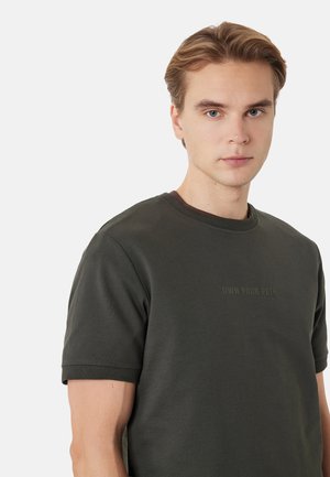 Junger Mann mit hellbraunen Haaren, der ein dunkelgrünes Kurzarmhemd mit dem Schriftzug "OWN YOUR PATH" auf der Brust trägt, blickt nach vorne.