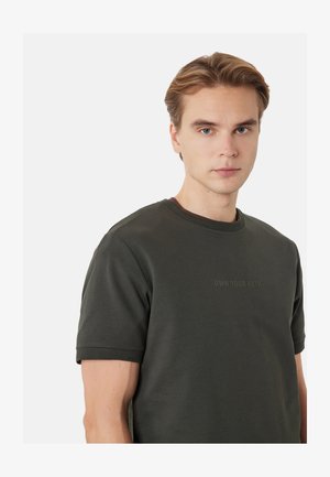 Jeune homme aux cheveux châtain clair portant une chemise verte foncée à manches courtes avec le texte «OWN YOUR PATH» sur la poitrine, regardant vers l'avant.