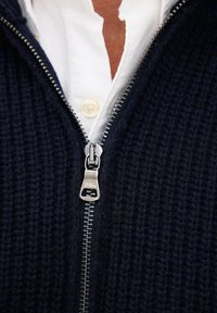 Primo piano di una zip in metallo argentato su un indumento in maglia nera dalla texture accentuata sopra un colletto di camicia bianca.