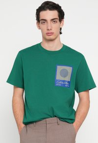 Zelené bavlněné tričko s krátkými rukávy. Má přední kapsu s modrým obdélníkem a kruhovým obrázkem, doplněné textovým detailem.