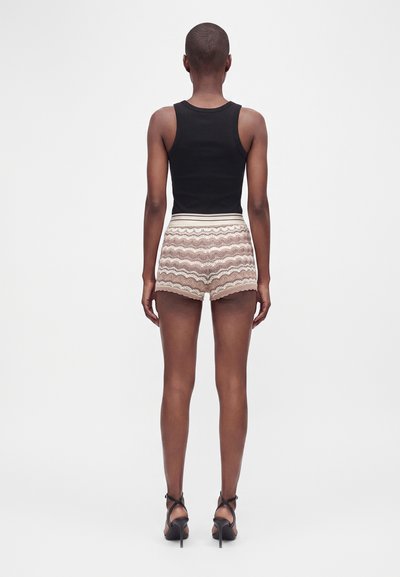 Personne debout de dos, vêtue d'un haut noir sans manches, d'un short beige à motifs et de sandales noires à talons hauts.
