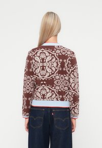 Braun gemusterter Pullover mit einem komplexen Design, gerippten, hellblauen Bündchen und Saum sowie kontrastierenden roten Akzenten.